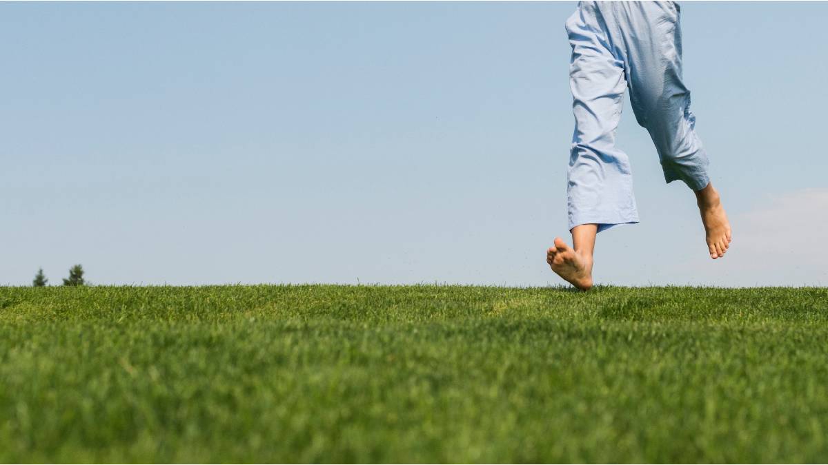 Persona caminando descalza sobre césped verde con pantalones claros, representando la sensación natural y libre del calzado barefoot.