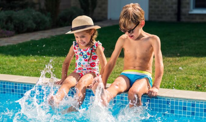 Niño y niña sentados al borde de una piscina en verano, chapoteando con los pies en el agua y sonriendo