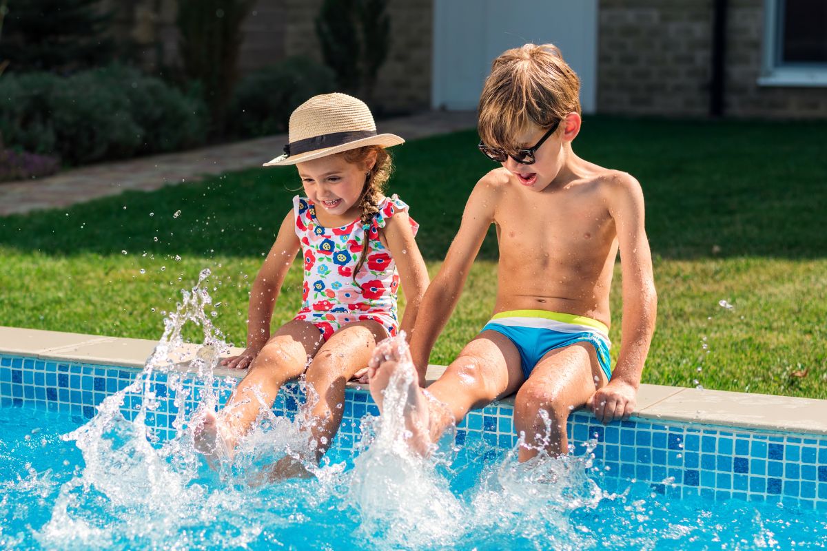 Niño y niña sentados al borde de una piscina en verano, chapoteando con los pies en el agua y sonriendo
